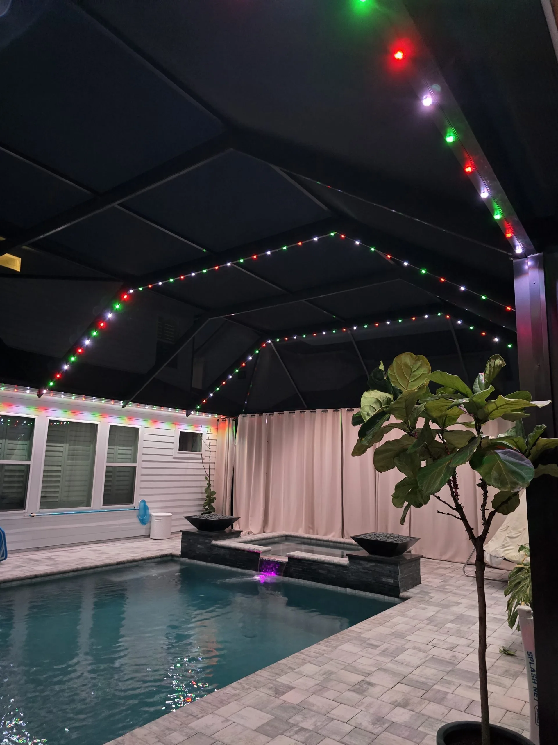 Pool cage lighting enhances a covered patio with colorful holiday lights beside a tranquil pool