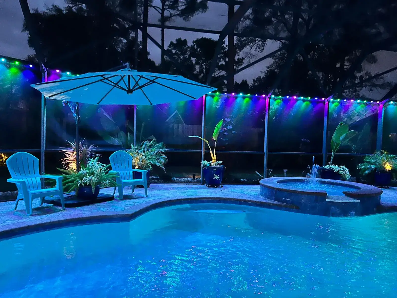 Brightly colored lights illuminate a serene pool area surrounded by plants and chairs